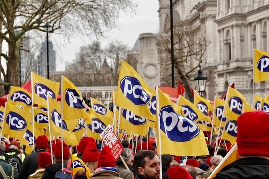 Crowd of protesters wearing red hats hold yellow "PCS" flags, standing near historic buildings.