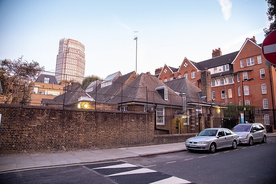 Street view of a residential area with school buildings, parked cars, and a tall tower in the background.