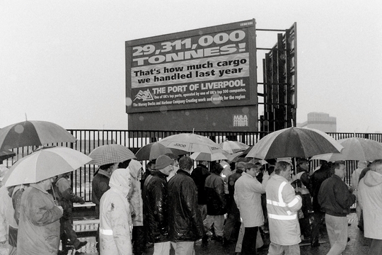 Umbrella-covered onlookers gather in the rain before a Port of Liverpool billboard claiming 29 million tonnes of cargo was handled last year, in monochrome.