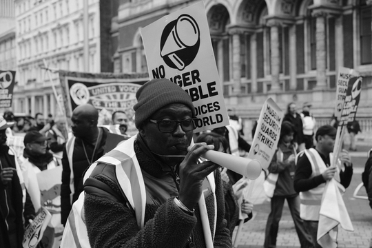 A outsourced security guard uses a vuvuzela on a protest during a joint strike at the Science Museum and Natural History Museum. In the background, other workers and supporters carry strike placards.