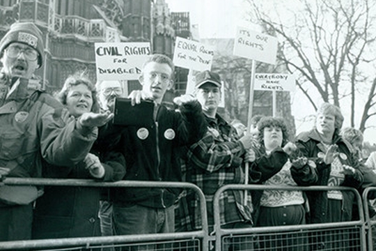 Protesters stand behind a barrier, holding signs advocating for disabled civil rights. Some of their hand positions indicate they are using sign language.