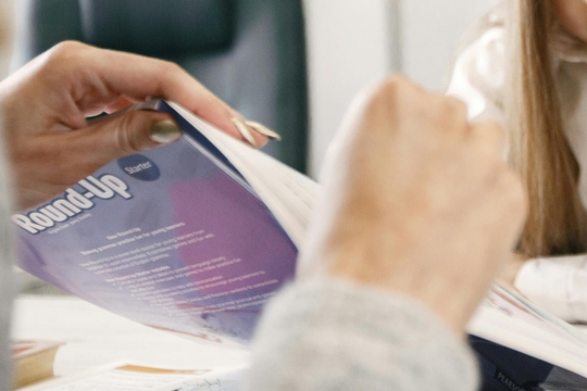 A close up picture of a teaching assistant holding a textbook up, whilst a student completes an exercise.